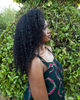 A woman with long, curly half wig black hair is shown in profile against a background of green foliage. She is wearing a dark green sleeveless top with a pink and white pattern.